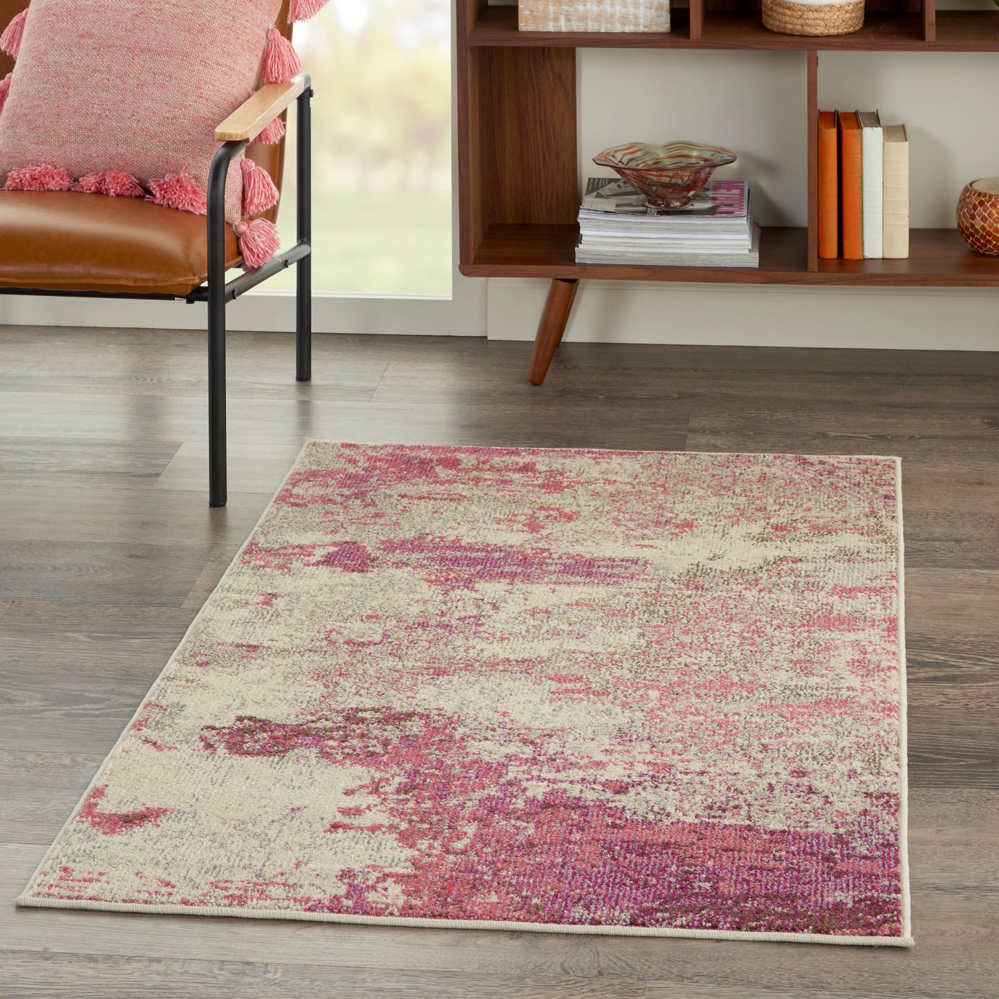 Rectangular cream area rug with pink abstract pattern on wood floor; complements mid-century bookshelf and pink chair.