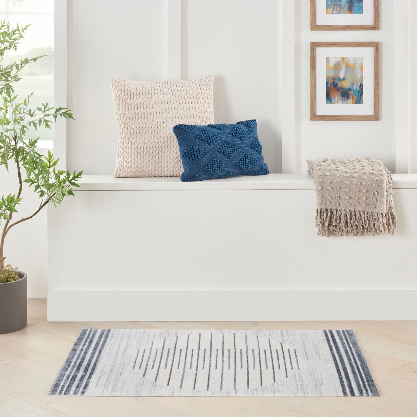 Blue-gray striped rug with a white built-in bench, beige knit cushion, blue diamond-knit pillow, beige fringe throw, and a plant.