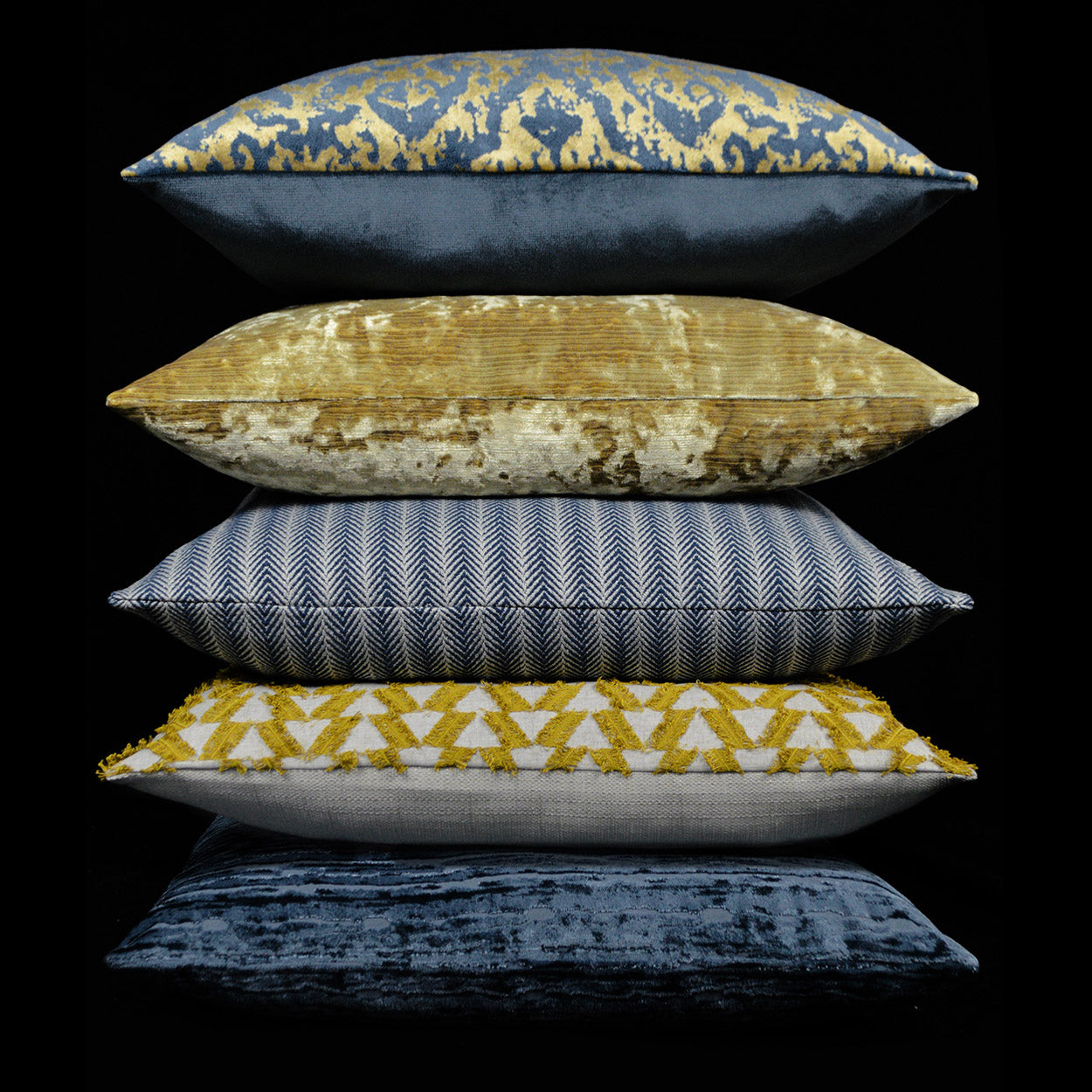 Five decorative throw pillows stacked: indigo with gold motif, gold velvet, blue herringbone, yellow triangles, navy velvet.