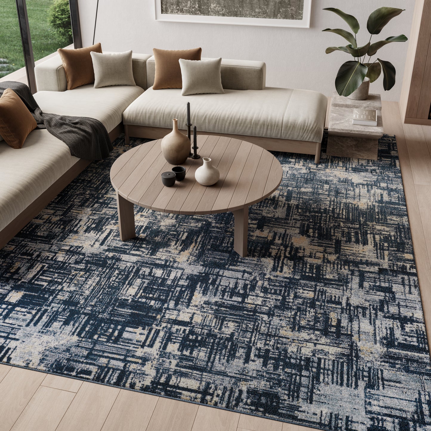 Blue patterned rug on floor with beige L-shaped sofa, brown and taupe cushions, a round wood coffee table, vases, candles, and a plant.