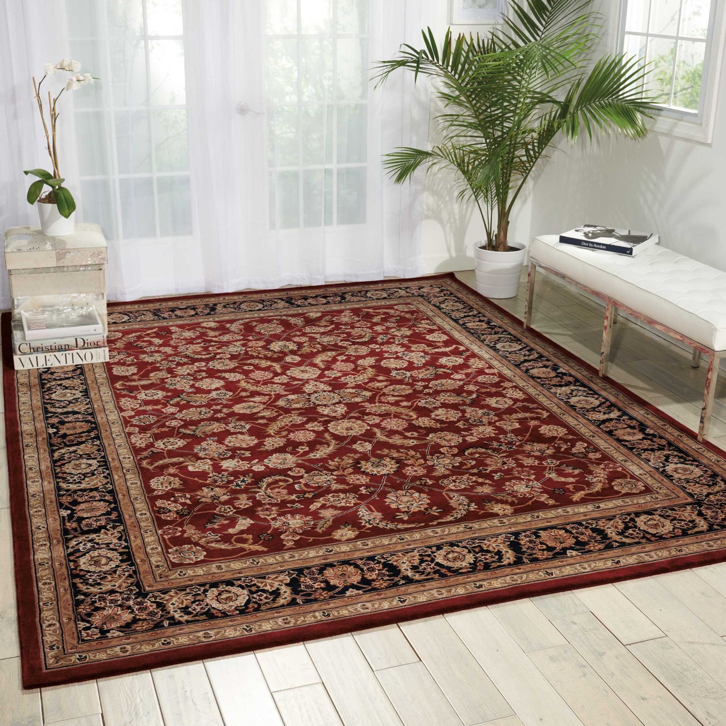 Large red oriental area rug with ornate floral border in a bright room; white tufted bench, palm plant, sheer curtains.