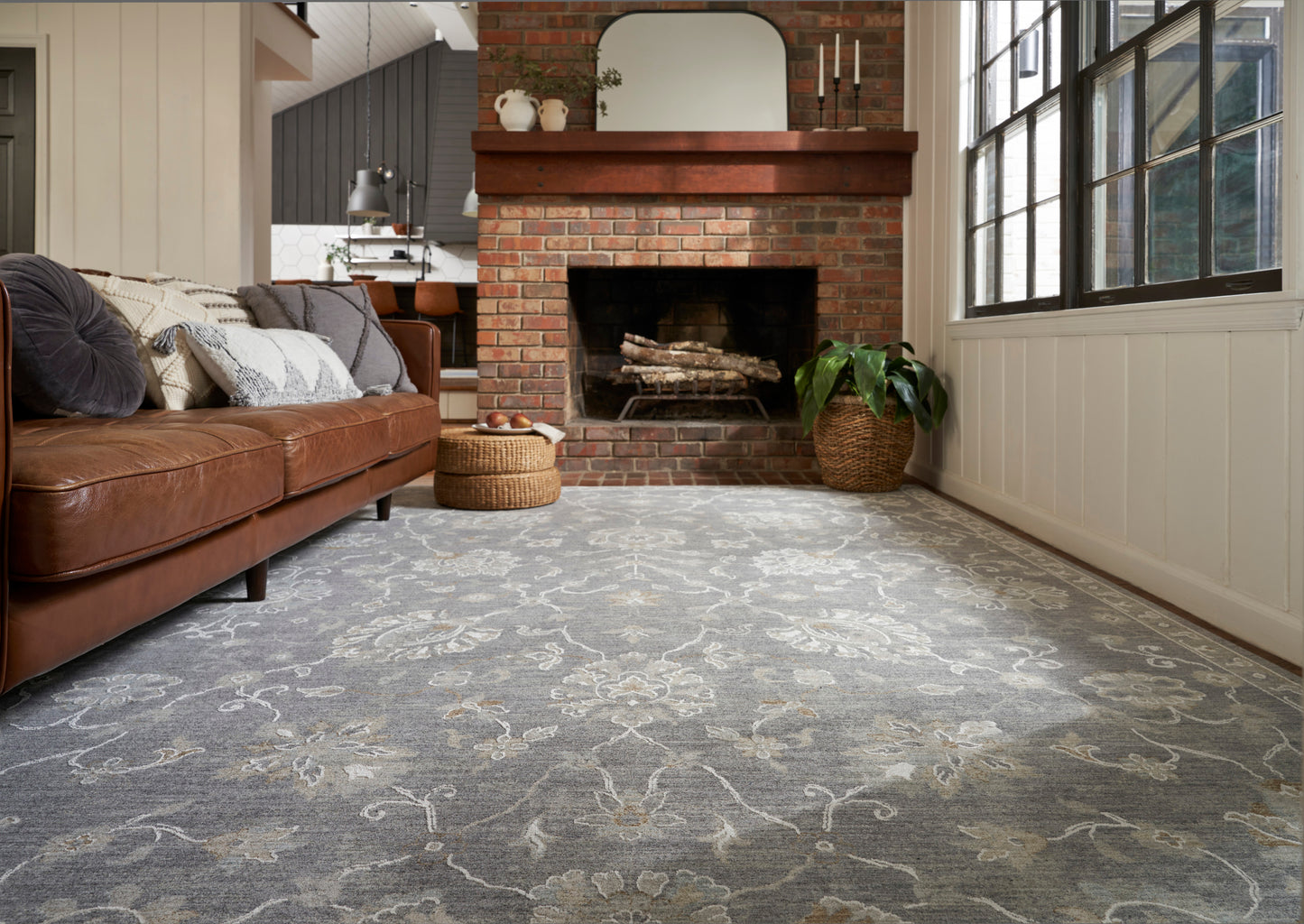 Cozy living room with a gray ornate-pattern area rug, brown leather sofa, brick fireplace, and large windows.