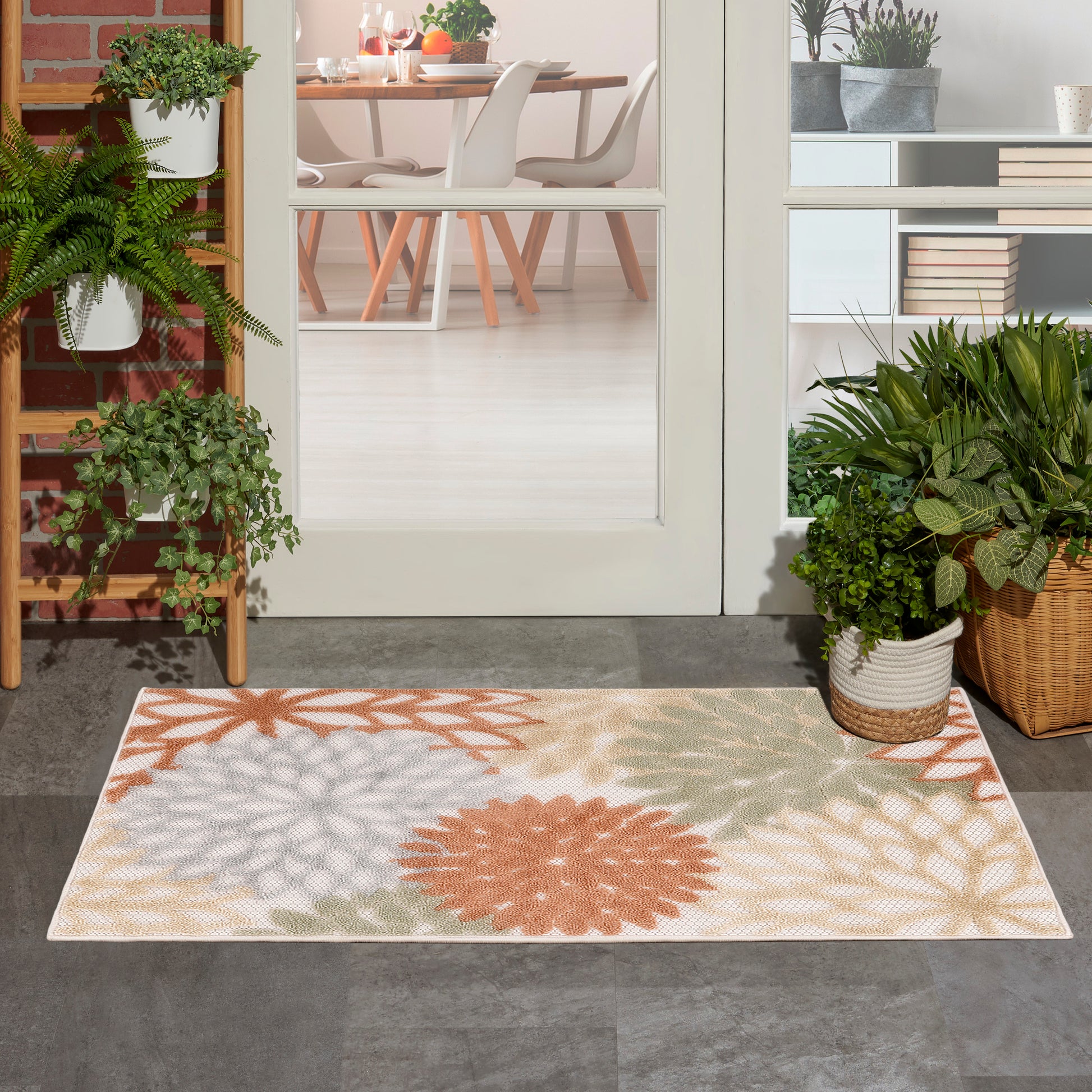 Outdoor rug with orange, peach and green floral pattern; glass door opens to a bright dining room with potted plants nearby.