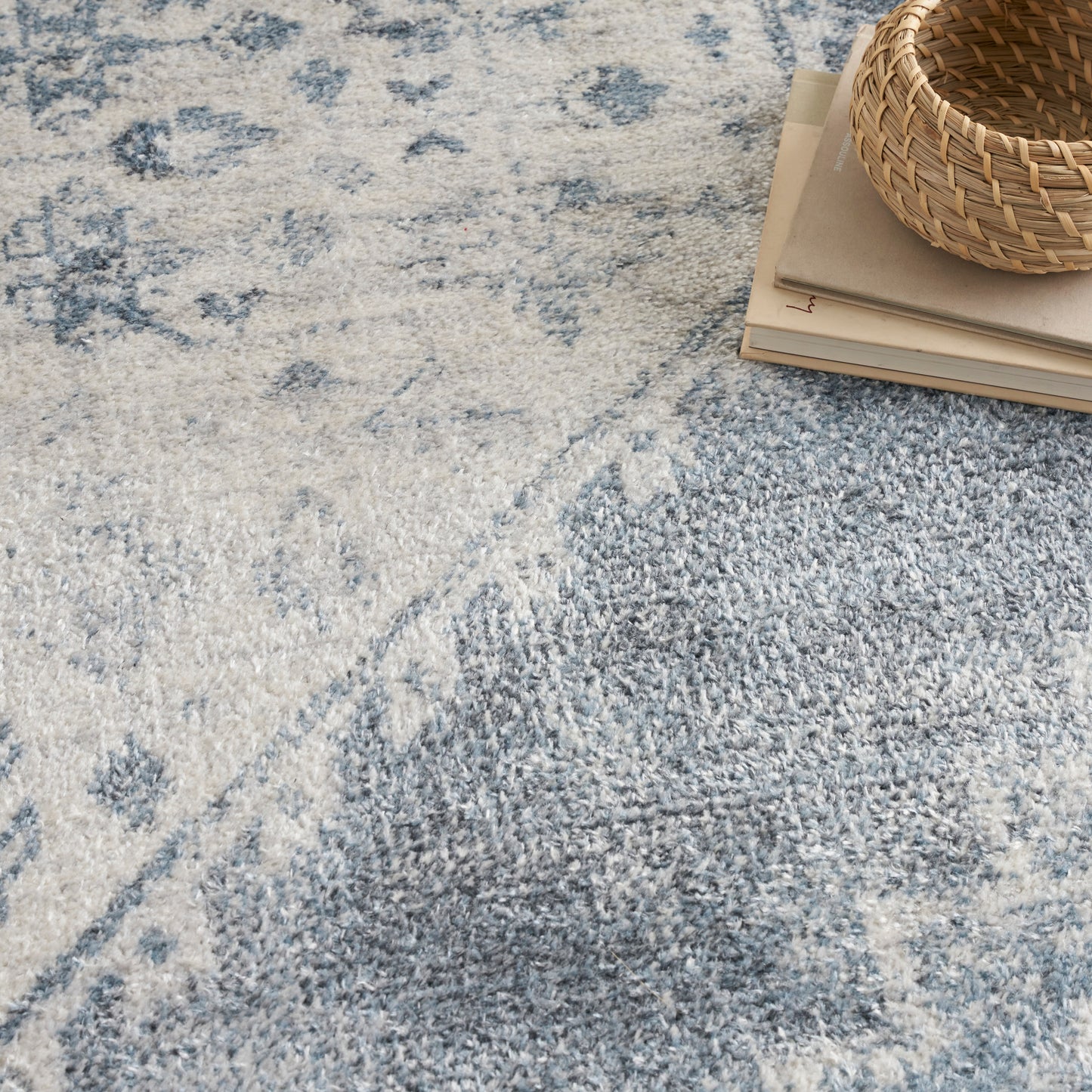 Blue & ivory patterned area rug with plush pile, stacked books & a woven basket in corner.