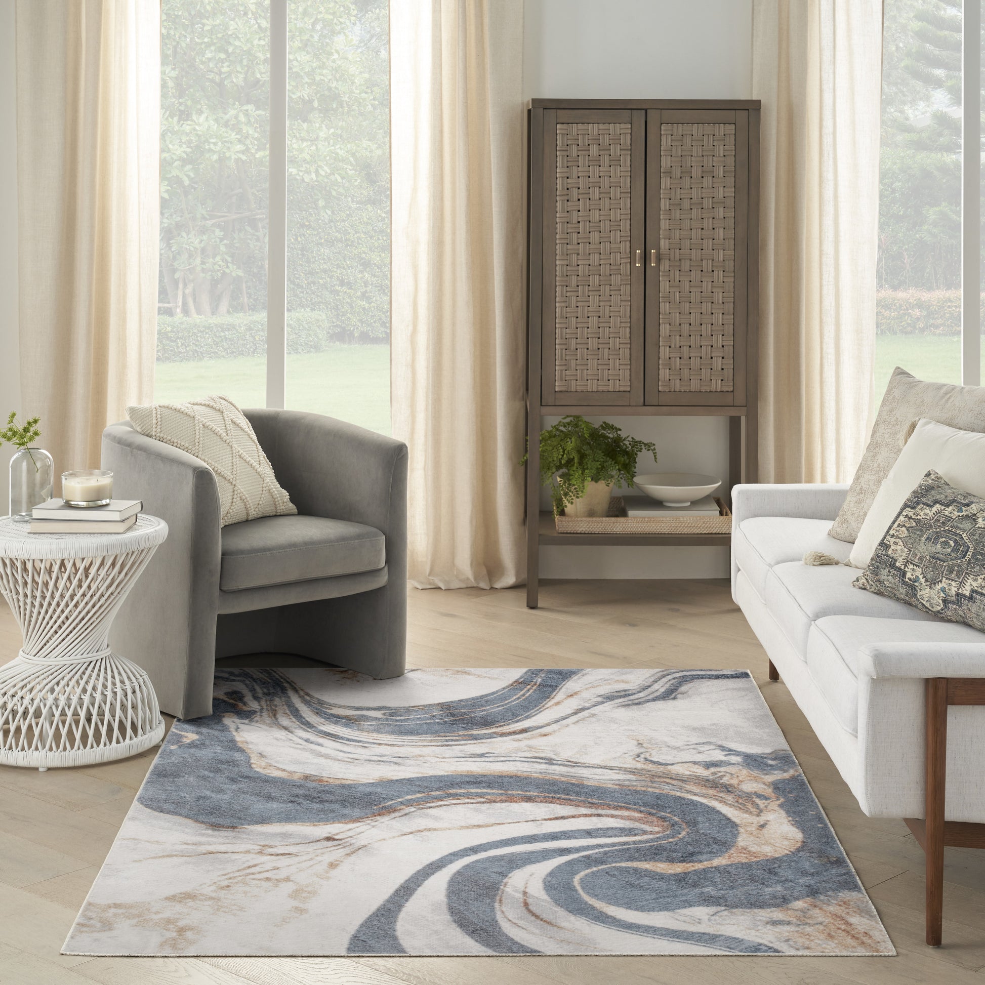 Bright living room with beige curtains, gray armchairs & white sofa, abstract blue-beige rug, wicker cabinet & plant.