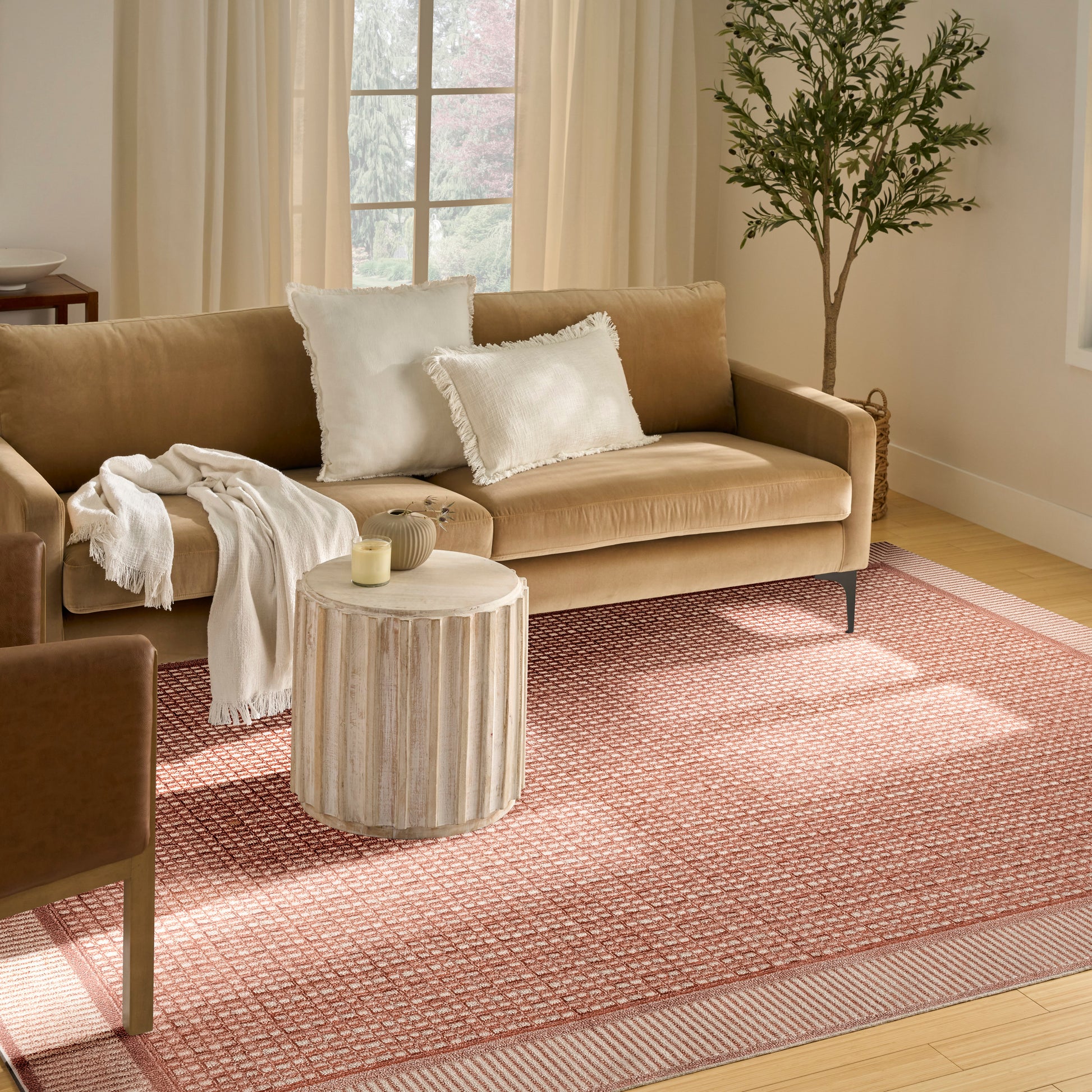 Beige fabric sofa with white fringe pillows and throw blanket, round light wood side table, pink rug, tall plant nearby.