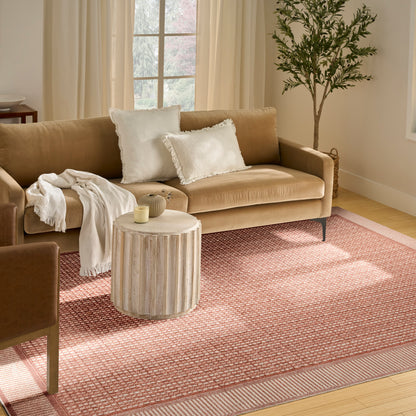 Beige fabric sofa with white fringe pillows and throw blanket, round light wood side table, pink rug, tall plant nearby.