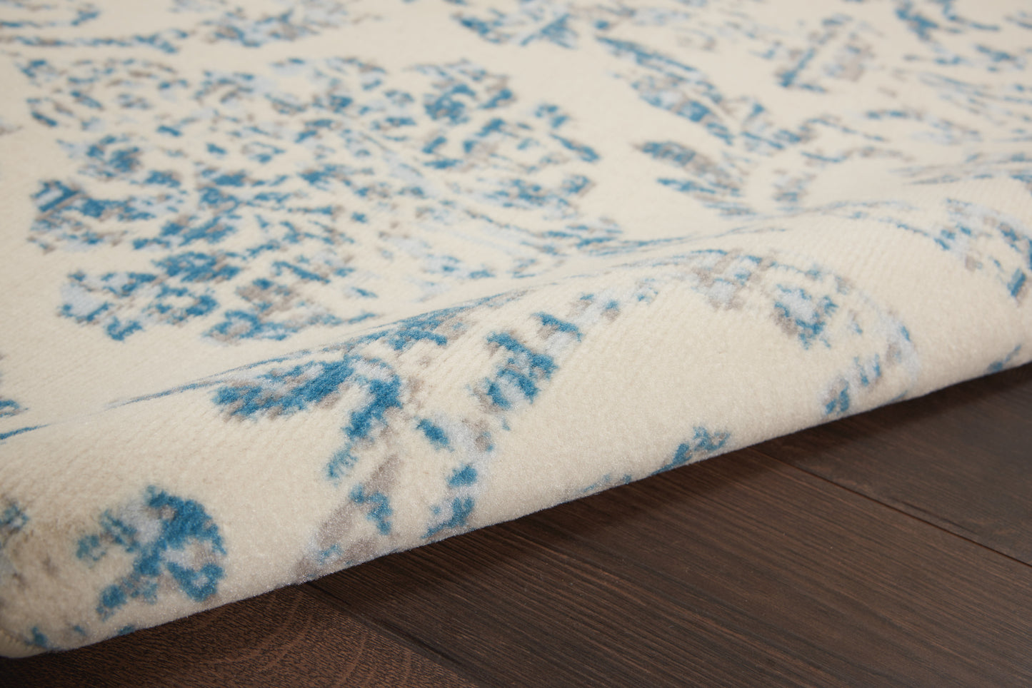Cream woven rug with blue botanical/abstract pattern, soft texture, draped on dark wood floor.