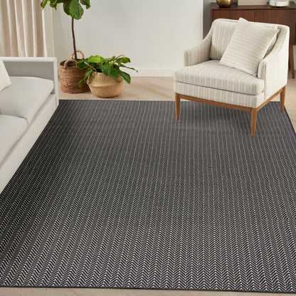 Dark herringbone area rug under a cream striped armchair with wooden legs; potted plants and a beige sofa nearby.