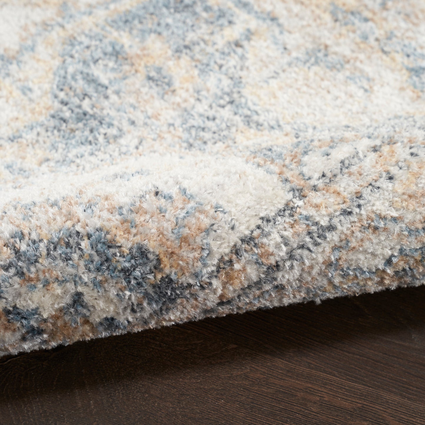 Close-up of a plush shag rug in blue, beige, gray and tan tones, edge resting on dark wood floor.