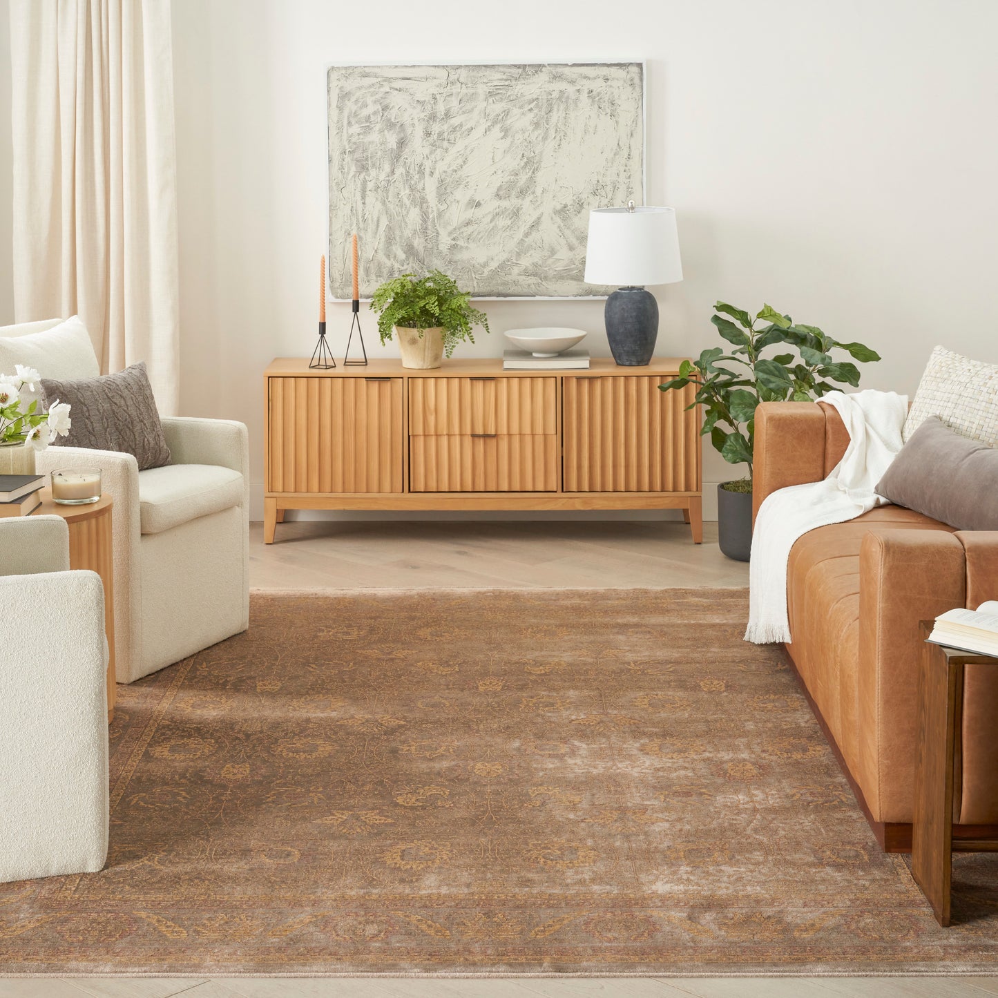 Cozy living room with a wood sideboard, beige armchairs, tan sofa, green plants, and abstract wall art over a patterned rug.