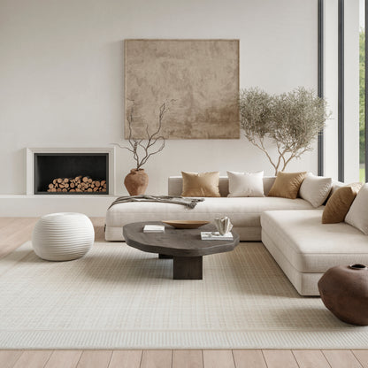 Neutral living room with beige sectional, dark round coffee table, white pouf, light rug, fireplace, and potted trees.
