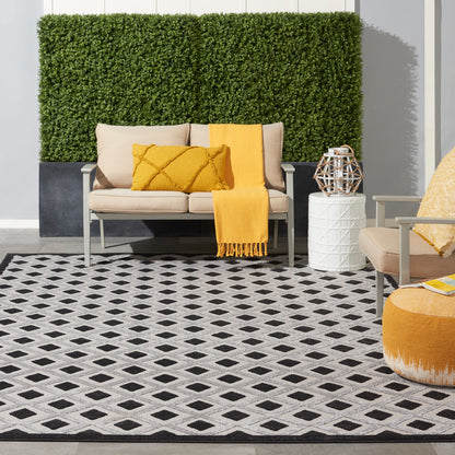 Outdoor beige sofa, mustard throw and pillows, black-and-white diamond rug, white side table with lantern, greenery wall.
