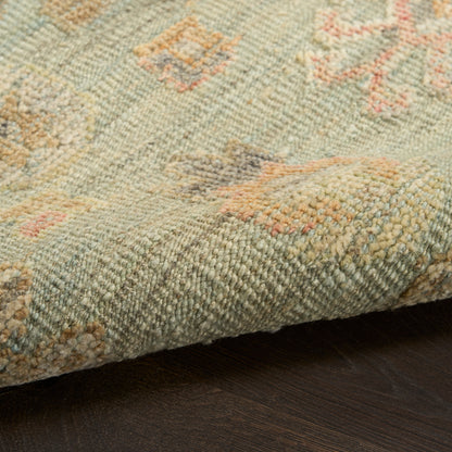 Pastel green oriental rug close-up with peach and pink motifs, textured knotted pile, folded corner on dark wooden floor.