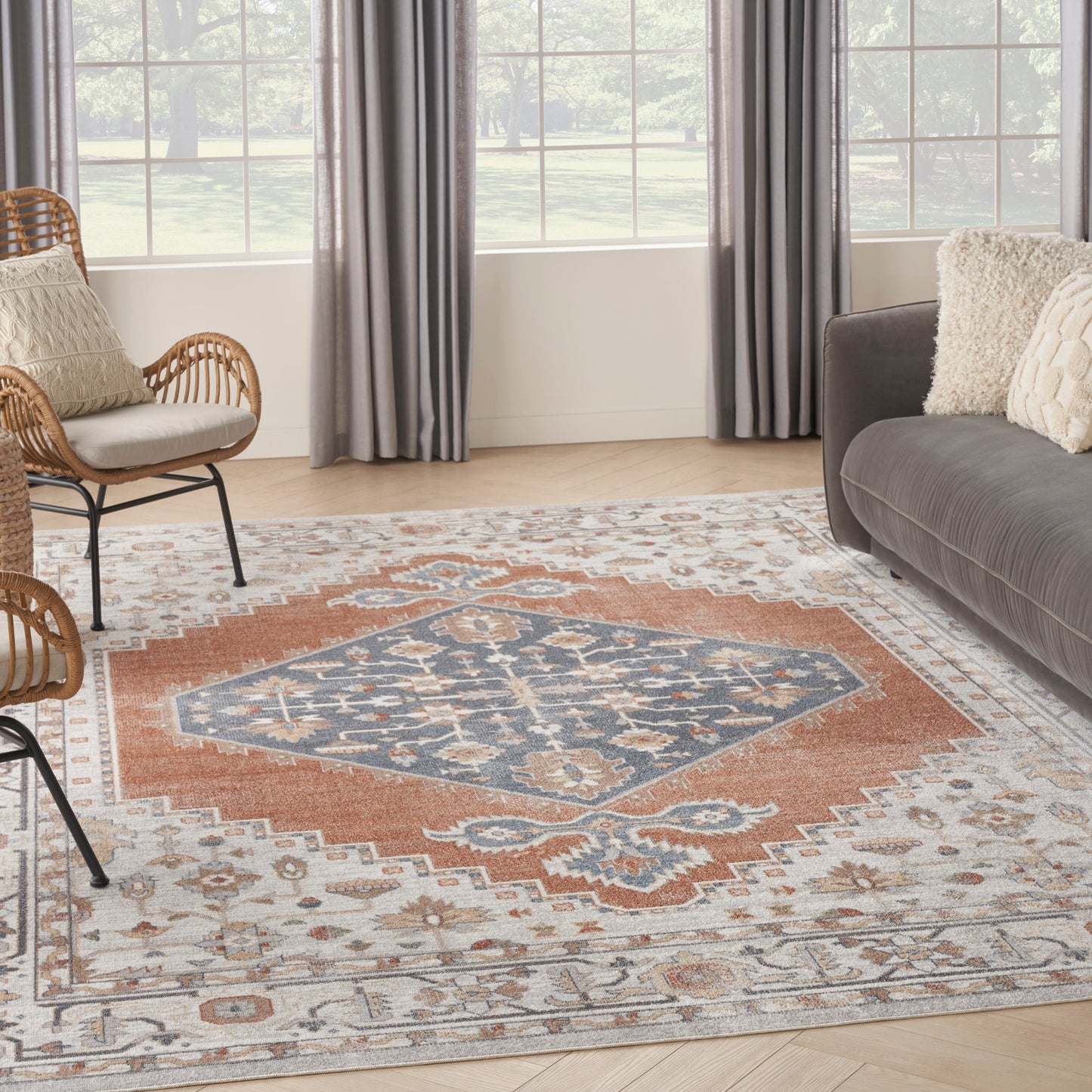Living room with rust-blue geometric area rug, gray sofa, wicker chairs, beige pillows, light wood floor and large windows.