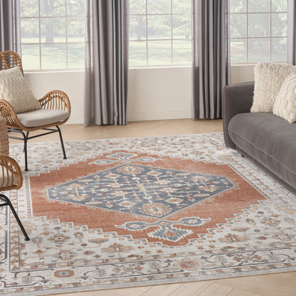 Living room with rust-blue geometric area rug, gray sofa, wicker chairs, beige pillows, light wood floor and large windows.