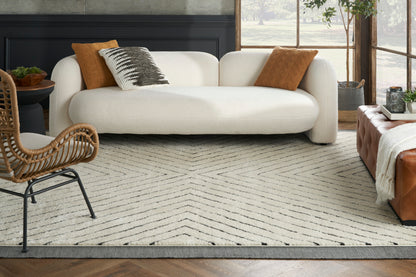 Cream curved sofa with orange and black/white patterned pillows, rattan chair, brown tufted ottoman, beige chevron rug.