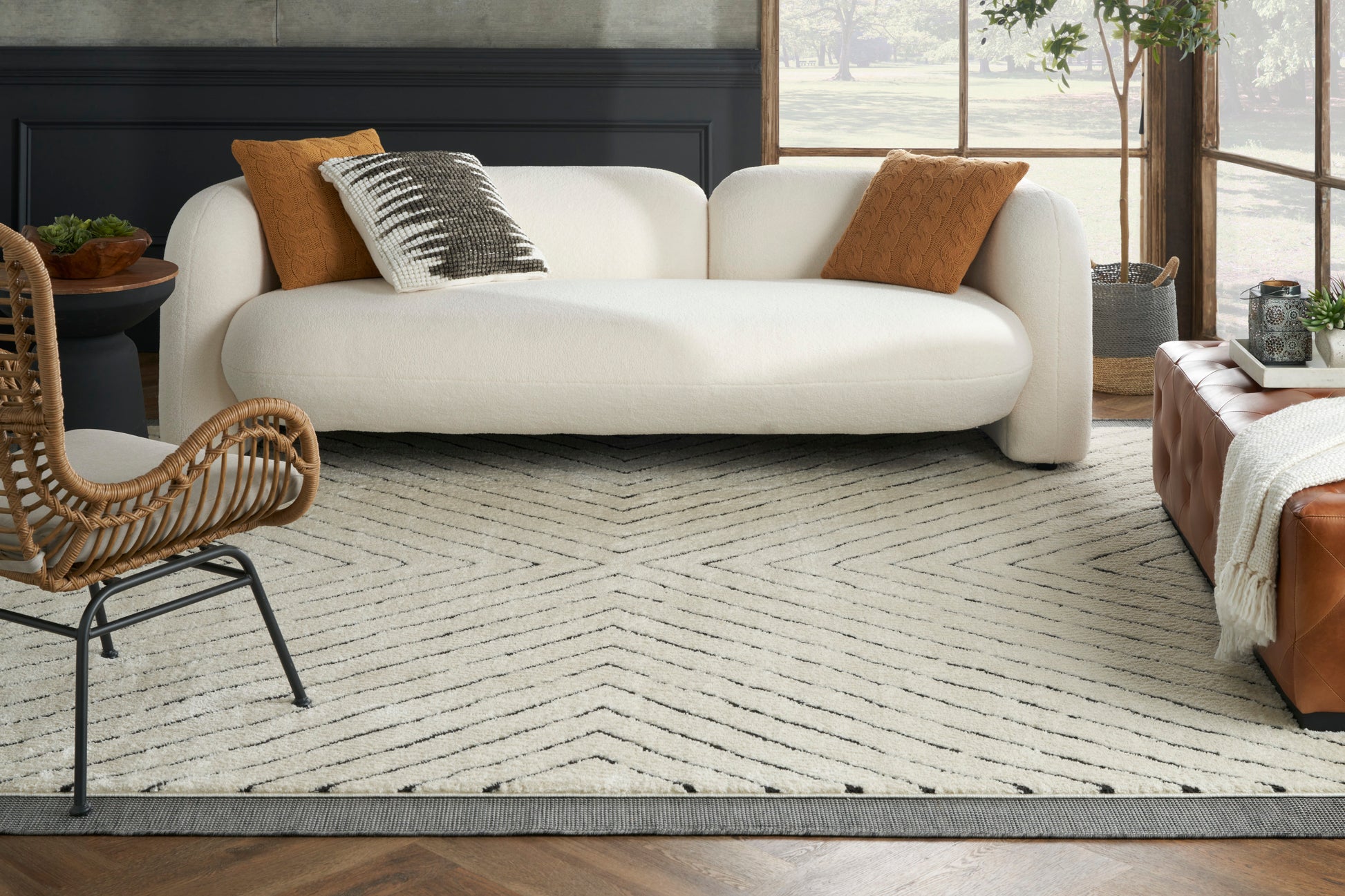 Cream curved sofa with rust pillows, woven rattan chair, chevron beige rug, brown leather ottoman in sunlit living room.