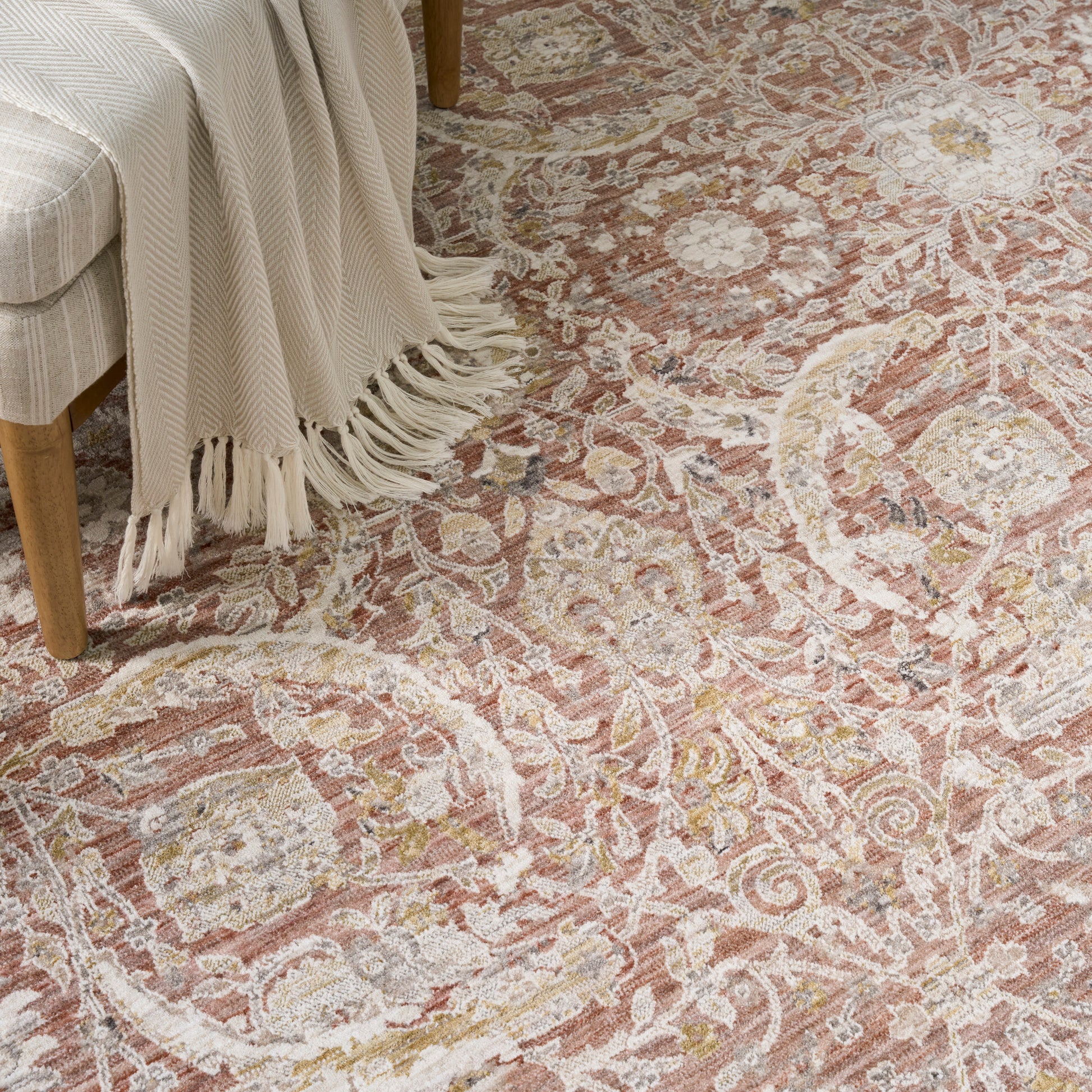 Intricate floral-scroll area rug in pink, cream and gold with a beige fringed throw over a light wood chair.