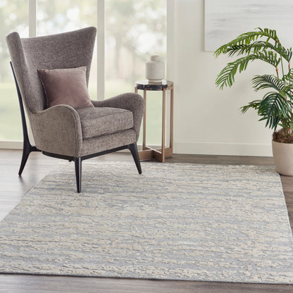 6' x 8' beige-gray rug in a living room, with a gray wingback chair with black legs, pink cushion, round side table, and a potted plant.