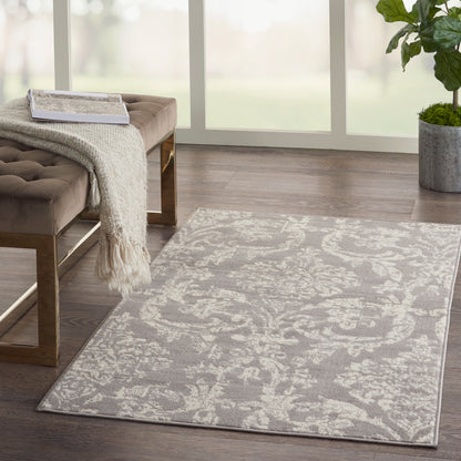 Light gray ornate area rug on wood floor beside a brown tufted bench with a cream throw, by a potted plant near large windows