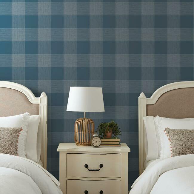Twin bedroom with navy plaid wallpaper, white nightstand, wicker lamp, clock, books, & plant.