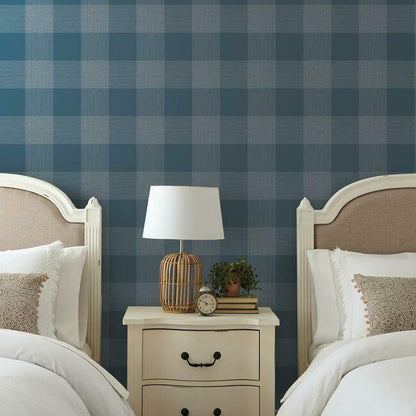 Twin bedroom with navy plaid wallpaper, white nightstand, wicker lamp, clock, books, & plant.