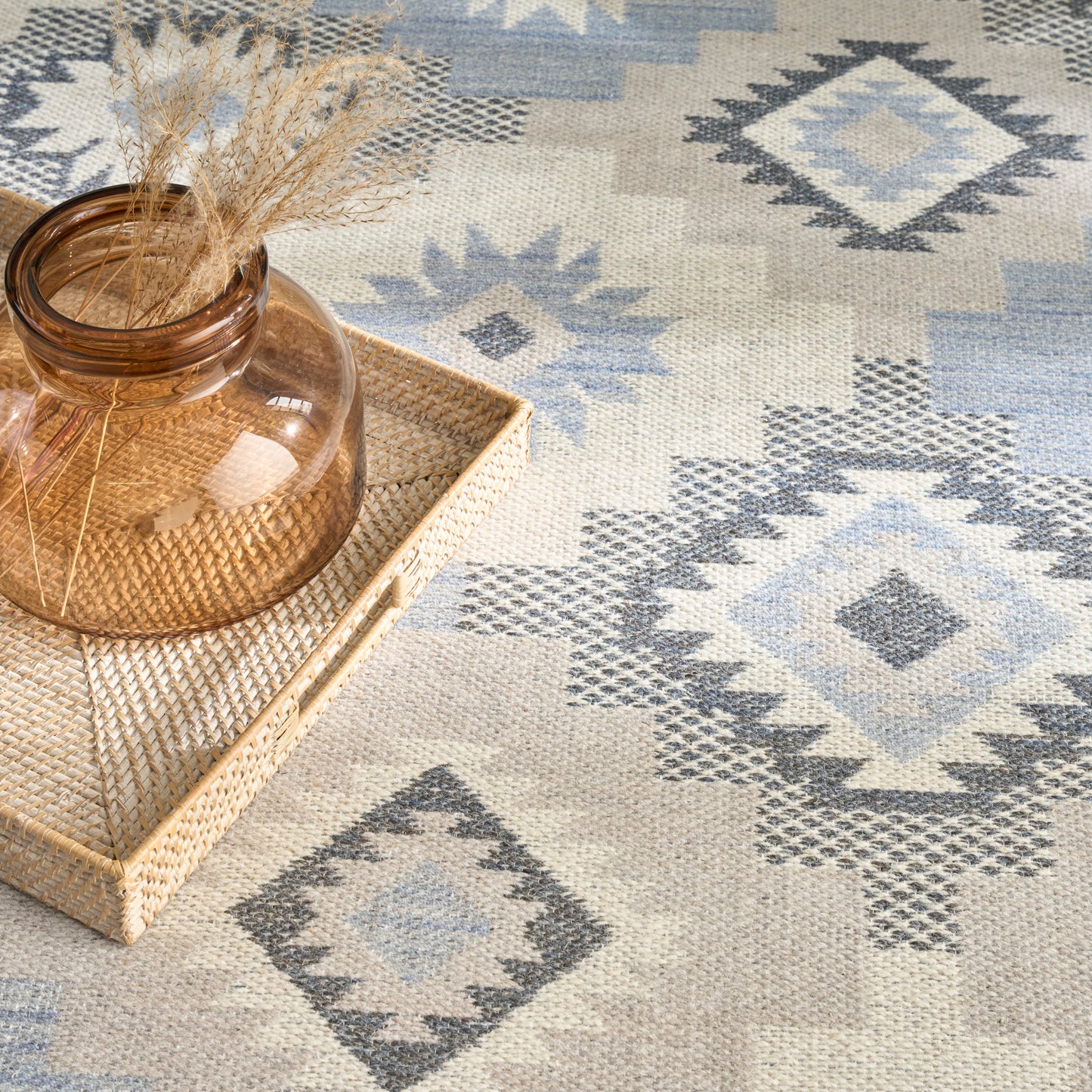 Amber glass vase with dried grasses on a woven rattan tray, set on a blue-gray diamond-pattern rug.