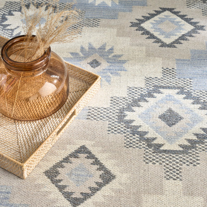 Amber glass vase with dried grasses on a woven rattan tray, set on a blue-gray diamond-pattern rug.