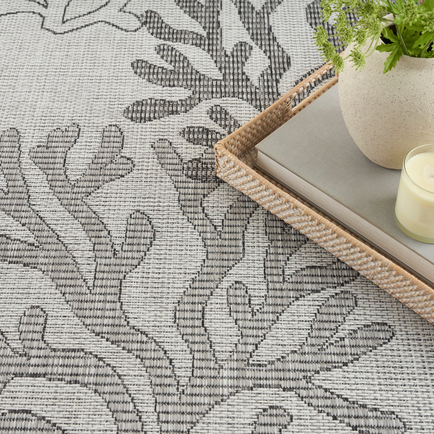 Grey patterned rug with abstract tree-branch motif; natural woven rattan tray holds a book, plant and candle.