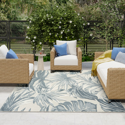 Outdoor wicker seating set with white cushions, blue accent pillows, palm-leaf rug, round side table, lush garden backdrop.