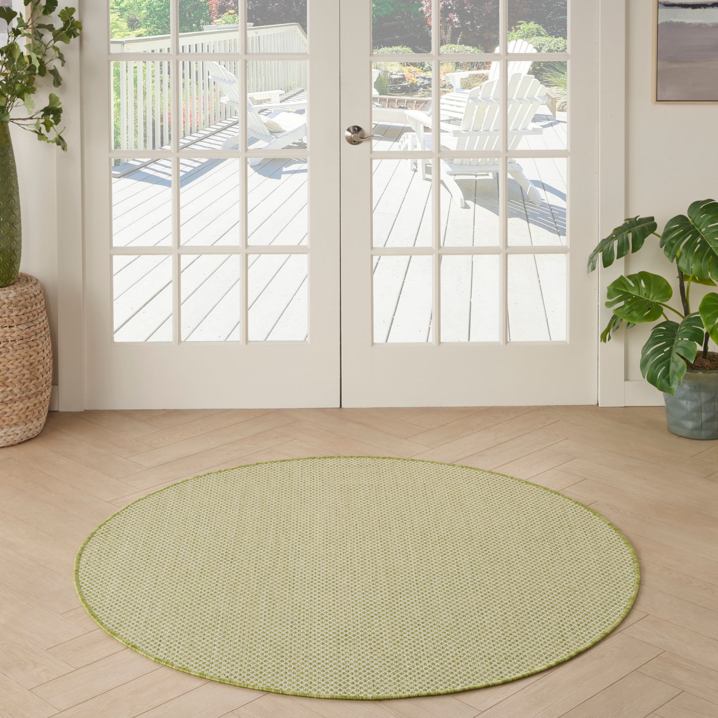 Round sage-green woven rug on light wood floor in a bright room with French doors and potted plants.
