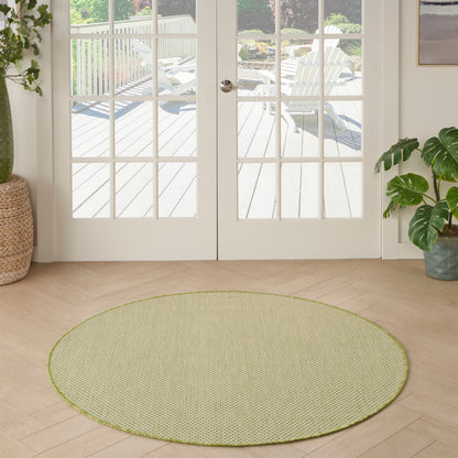 Round sage-green woven rug on light wood floor in a bright room with French doors and potted plants.