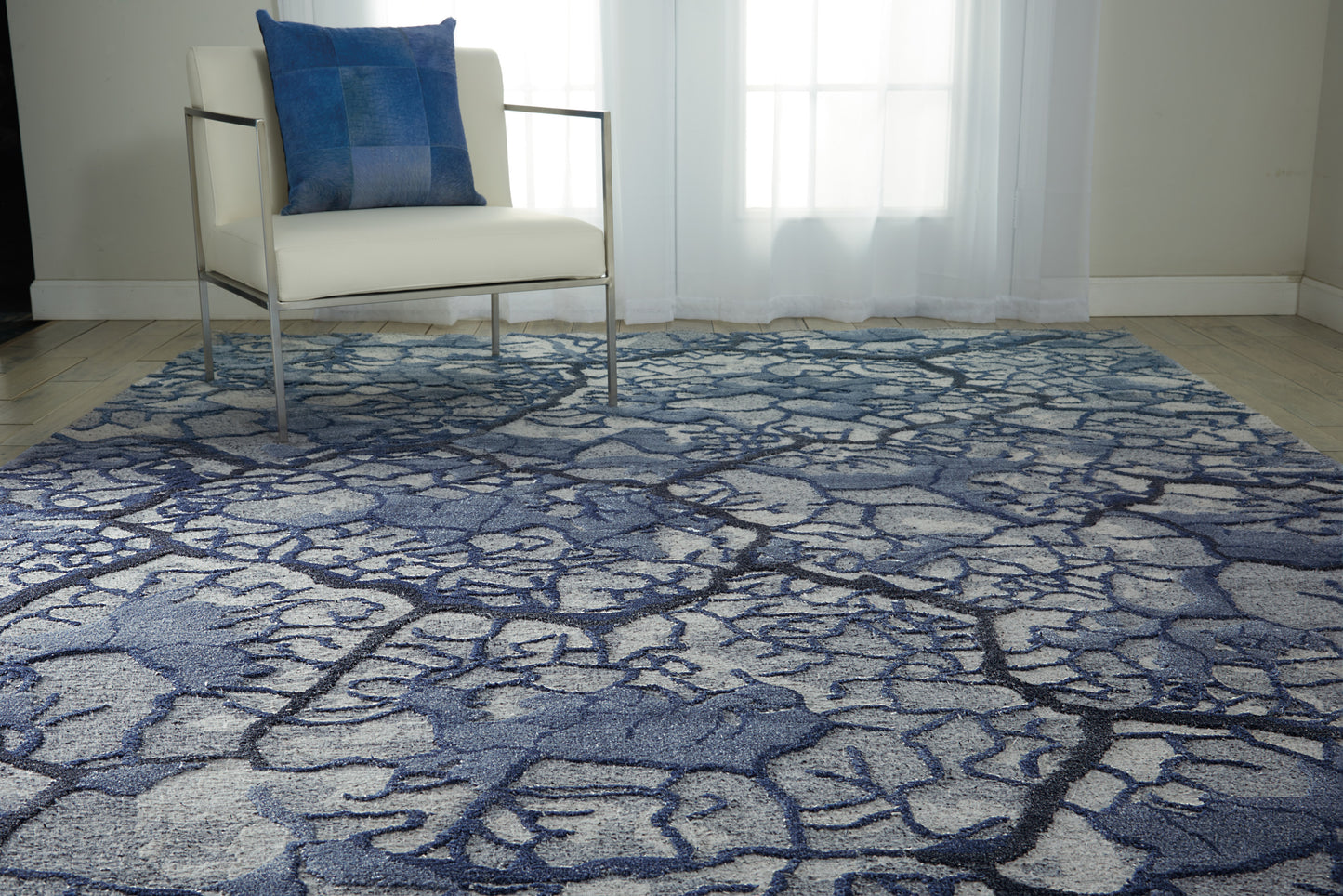 White modern armchair with chrome frame and blue pillow atop a blue-gray abstract patterned area rug in a bright room.