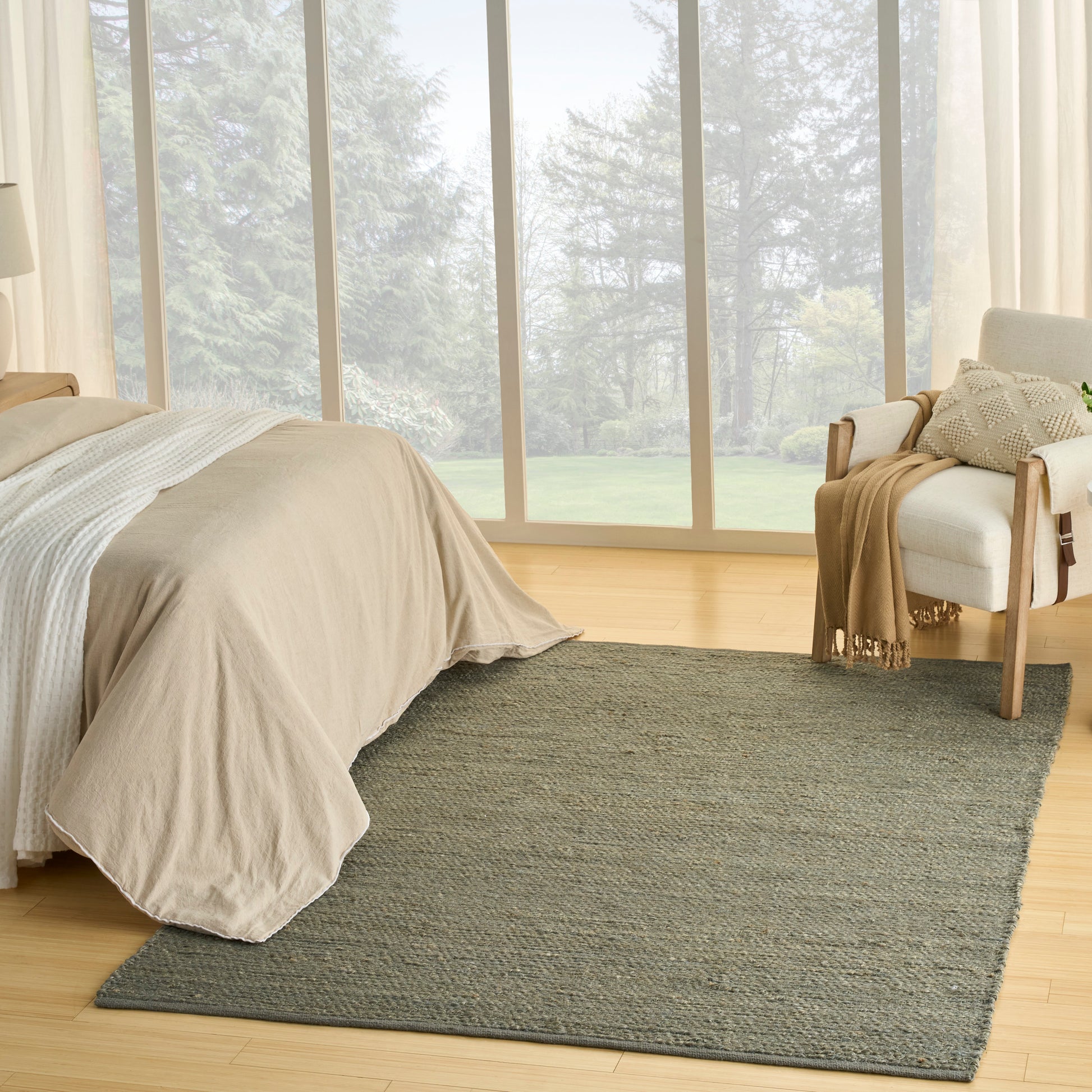 Neutral bedroom with beige duvet on a low bed, gray area rug, light wood floor, and a cream armchair near large windows.
