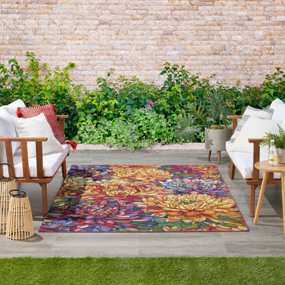 Outdoor patio with a vibrant floral rug, white cushioned wooden chairs, red pillows, lanterns, potted plants and stone wall.