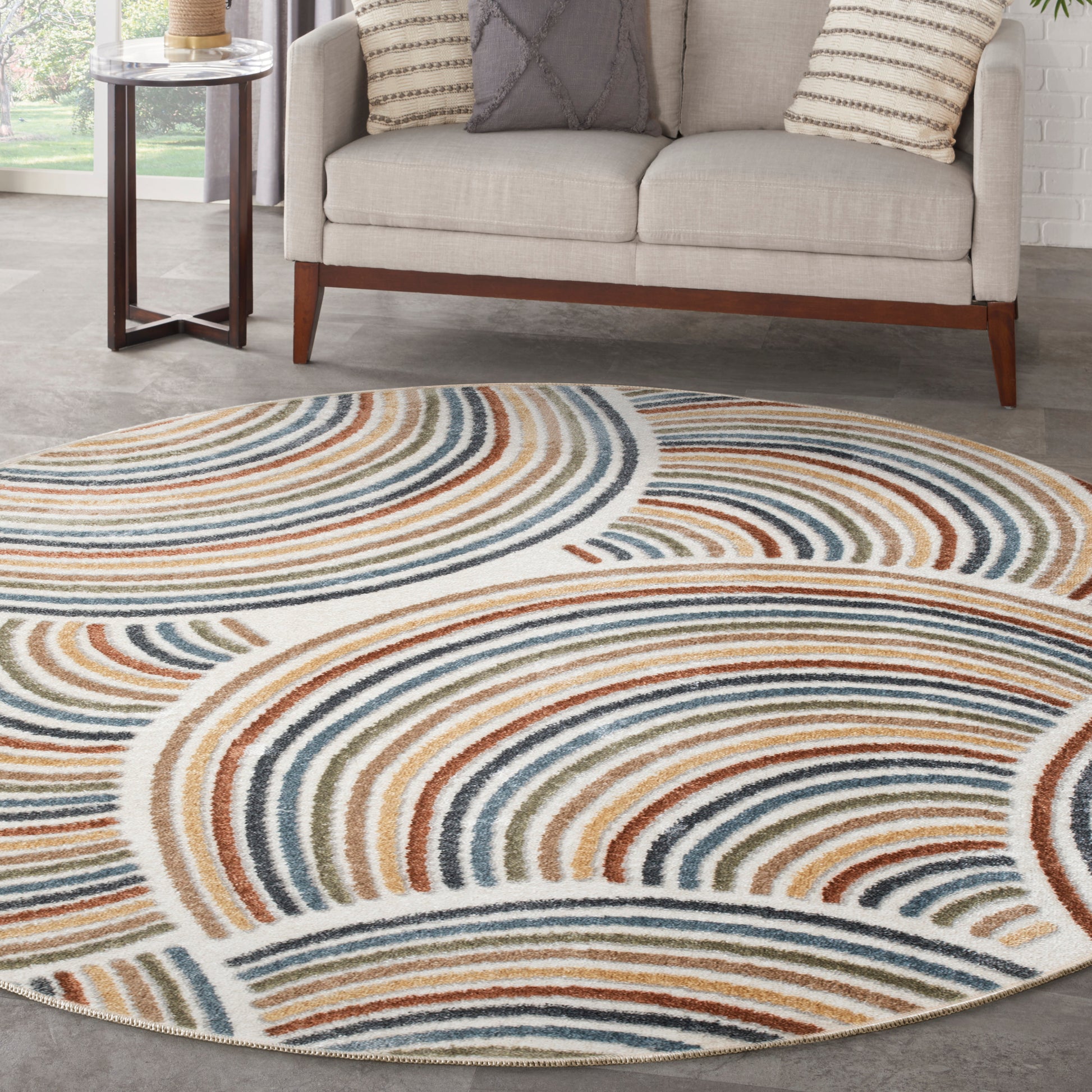 Circular area rug with concentric curved stripes in blue, teal, orange, tan on ivory; in a living room with a beige sofa.