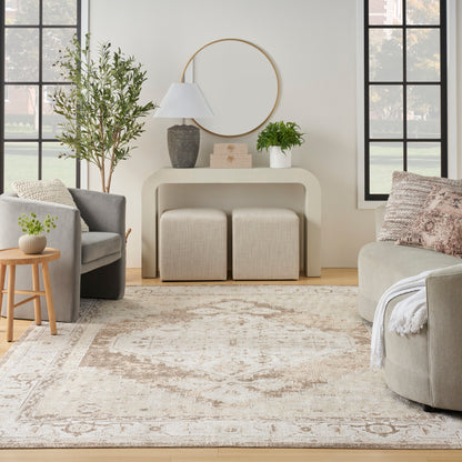 Neutral living room with curved beige console table, round mirror, potted plant, two knitted ottomans, and a patterned rug.