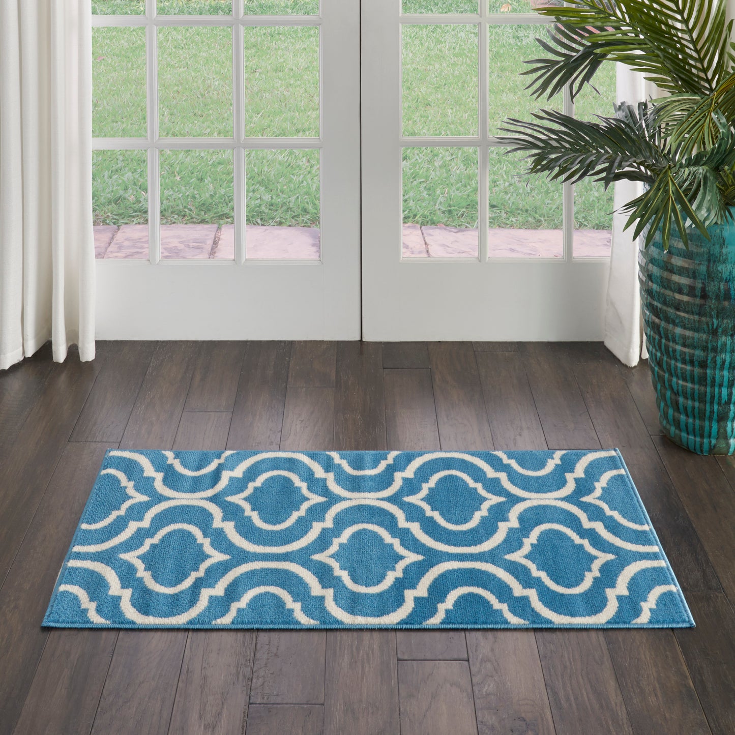 Blue rectangular area rug with white interlocking wave pattern in doorway on dark wood floor beside a potted plant.