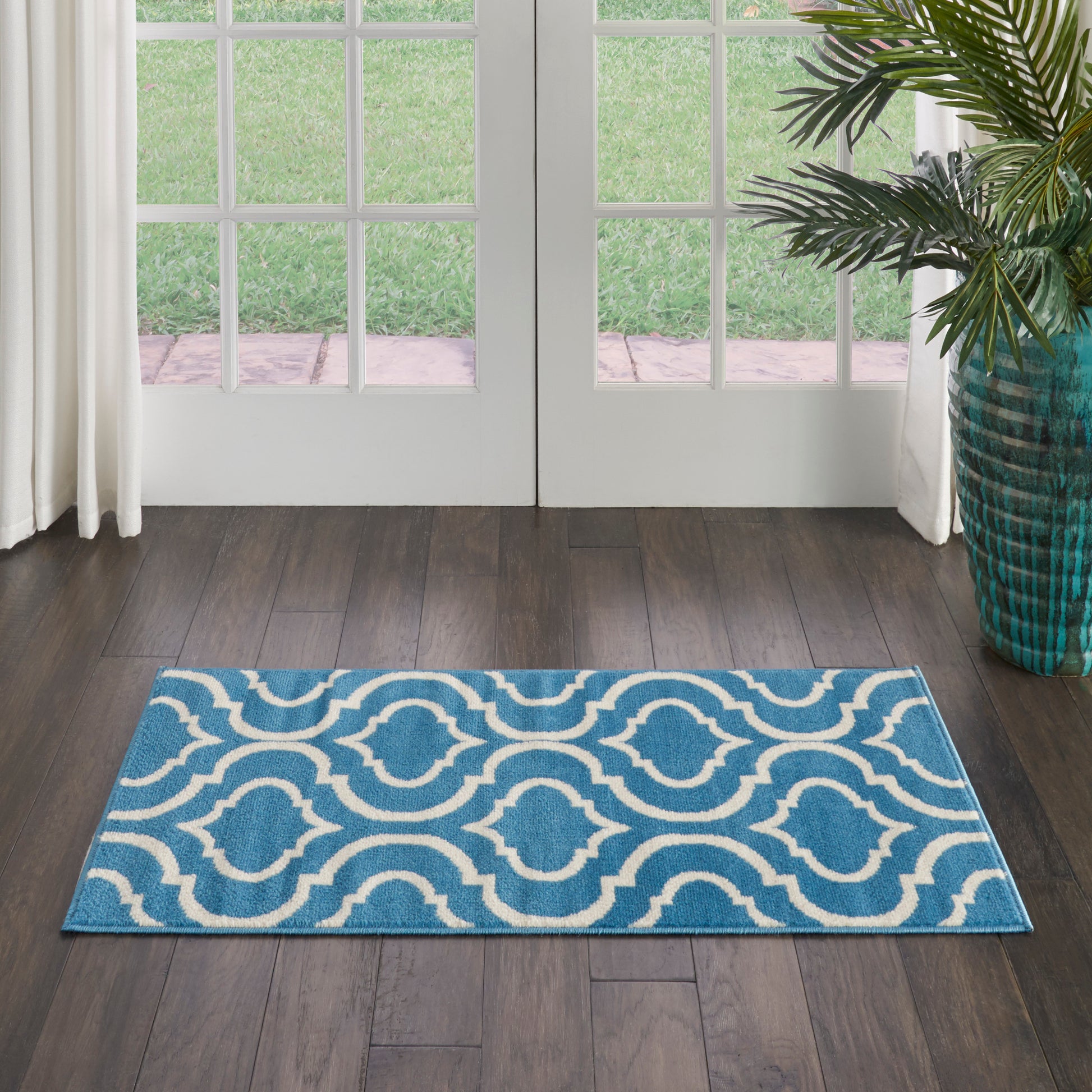 Blue rectangular area rug with white interlocking wave pattern in doorway on dark wood floor beside a potted plant.
