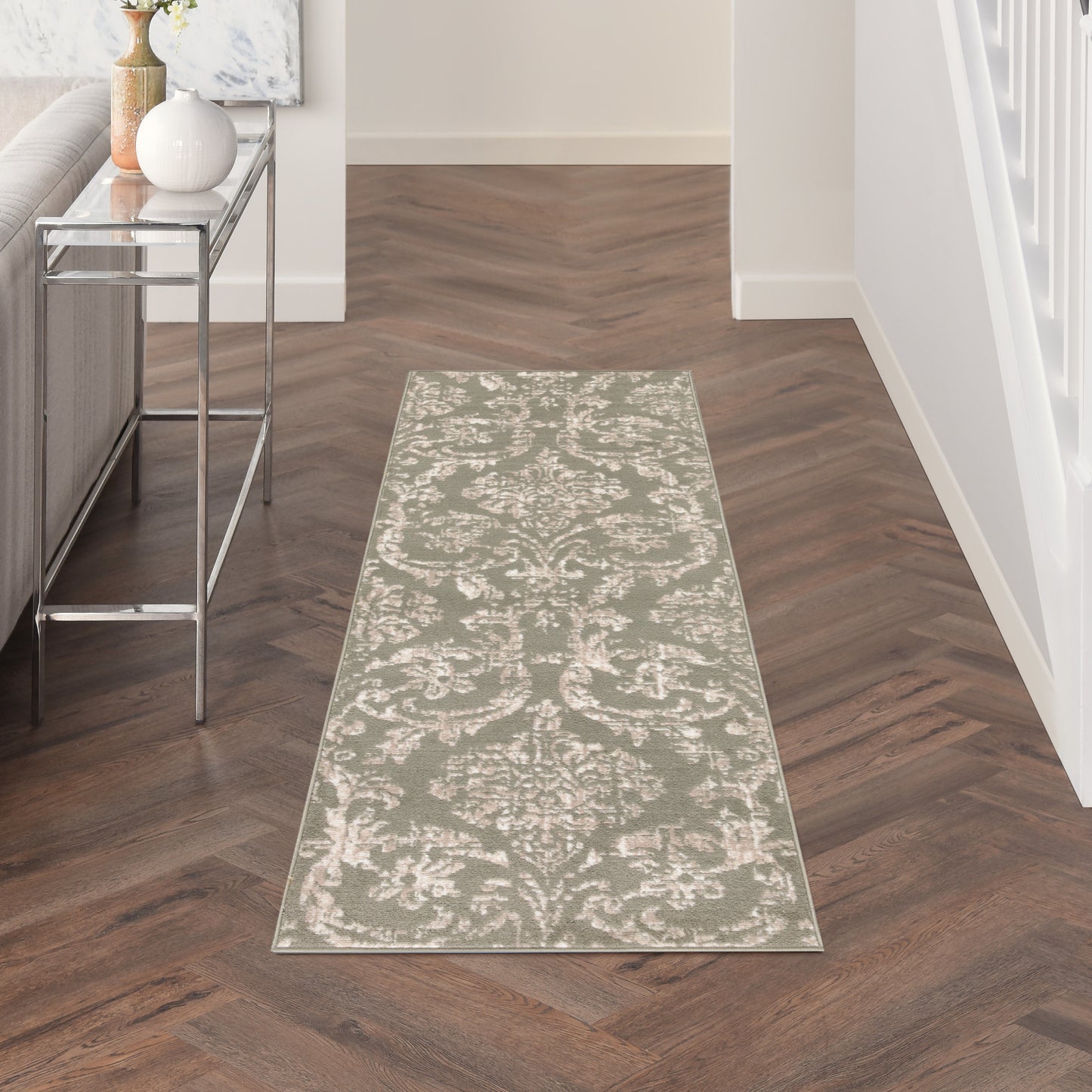 Long damask-patterned runner rug in pale sage and ivory on a dark wood hallway floor, beside a glass-metal console table.