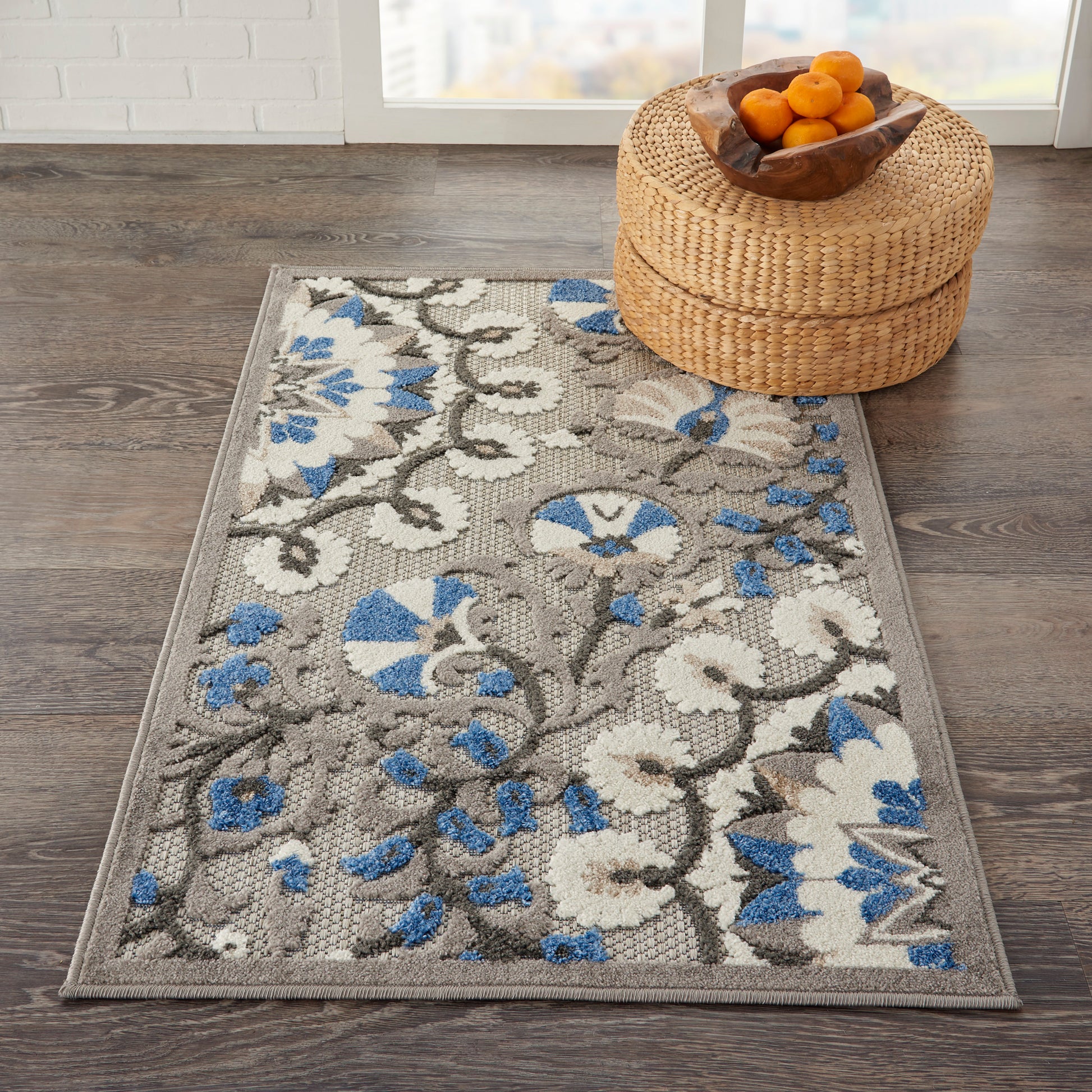 Gray floral area rug with white and blue blossoms on a wood floor, beside a woven round ottoman with a bowl of oranges.