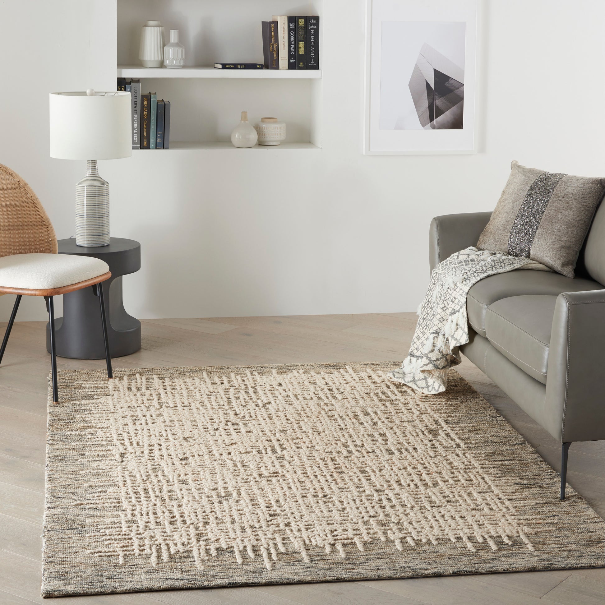 Neutral living room with beige woven rug, gray leather sofa, wicker chair, white shelves, and abstract wall art.