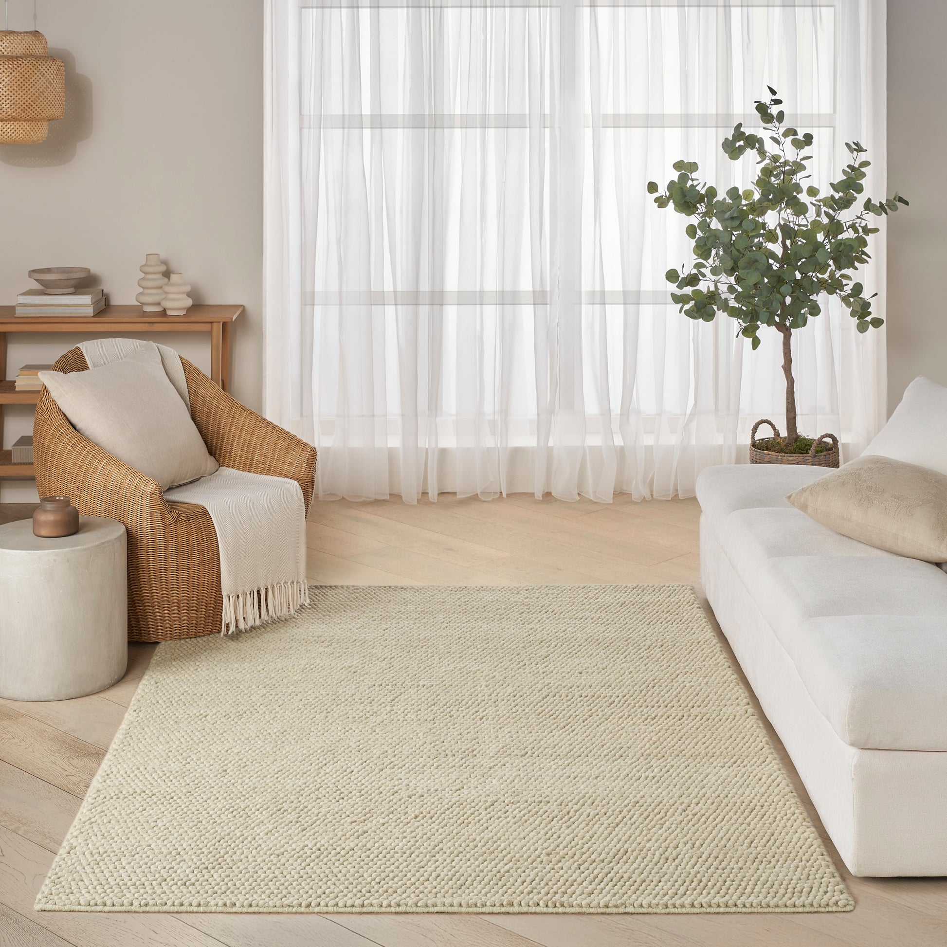 Neutral living room with white sofa, woven wicker armchair, beige jute rug, potted plant, sheer curtains, and wood tones.