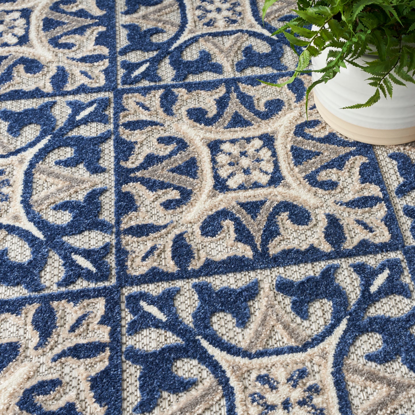 Blue and beige ornate patterned rug with arabesque motifs; woven texture; green potted plant in white pot at corner.