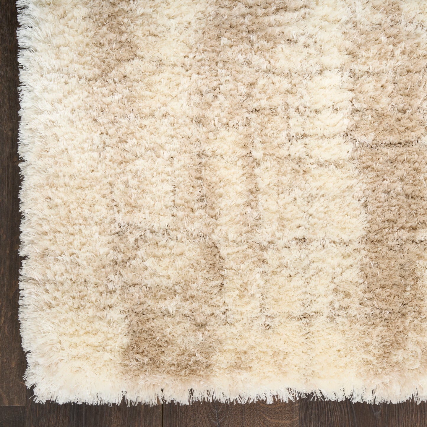 Beige shag rug with cream and tan tones, fluffy high-pile texture on dark wooden floor, fringe along edge.