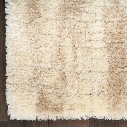 Beige shag rug with cream and tan tones, fluffy high-pile texture on dark wooden floor, fringe along edge.