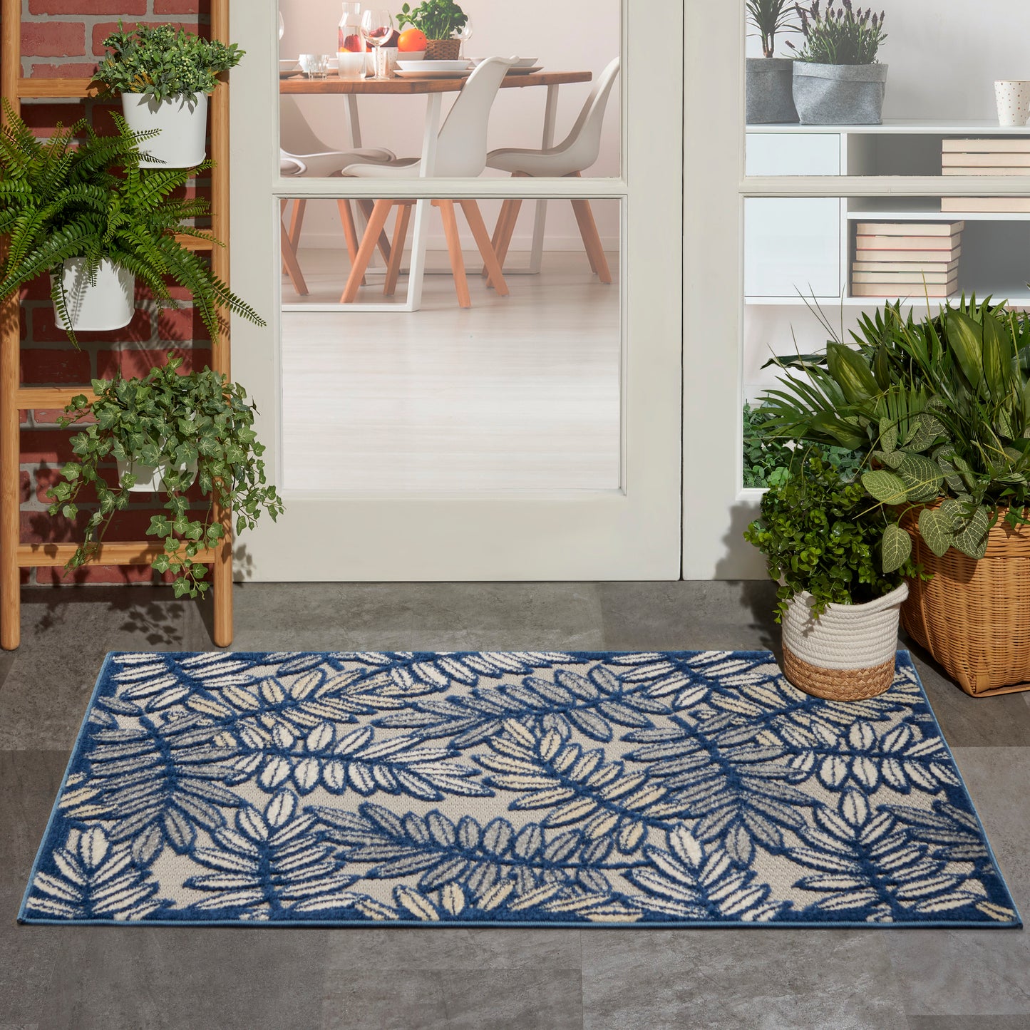 Blue and white fern-leaf patterned rug on a concrete entryway, flanked by plants, leading to a glass door and dining room.