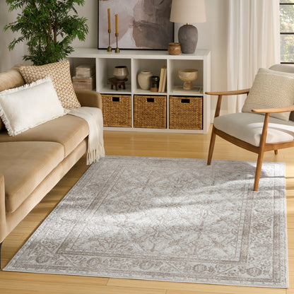 Light gray ornate border area rug on a light wood floor with beige sofa and wooden chair in a modern living room.