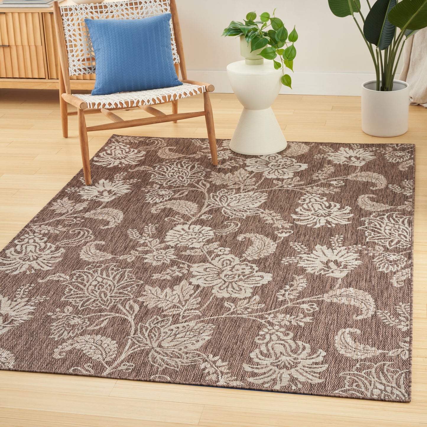 Brown floral-pattern area rug on light wood floor with a woven-seat wooden chair, blue cushion, white plant stand, and leafy potted plants.